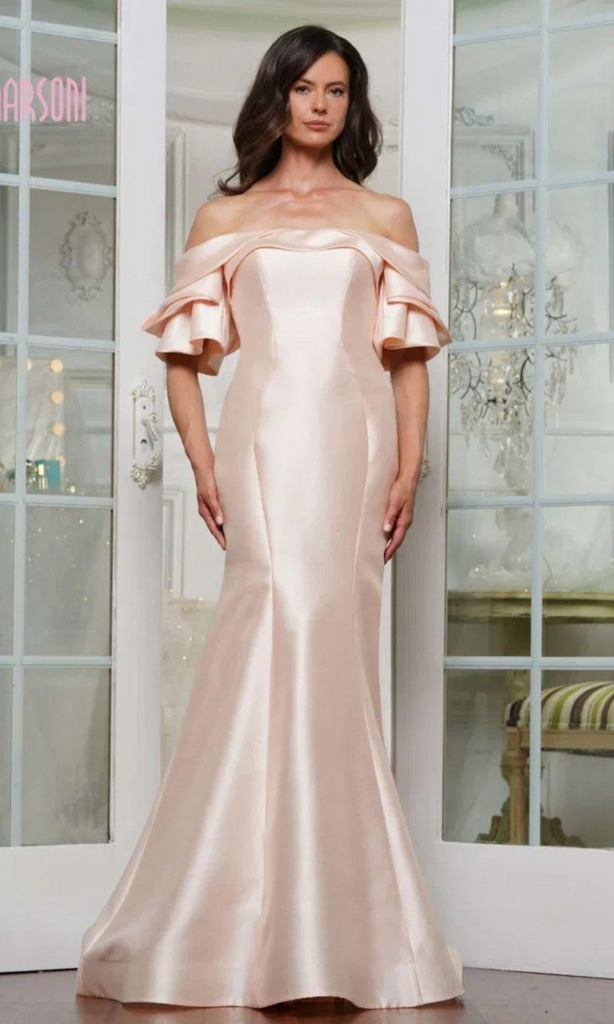 A woman stands indoors wearing the Marsoni MV1290, a long formal dress featuring off-the-shoulder ruffled short sleeves in light pink satin.